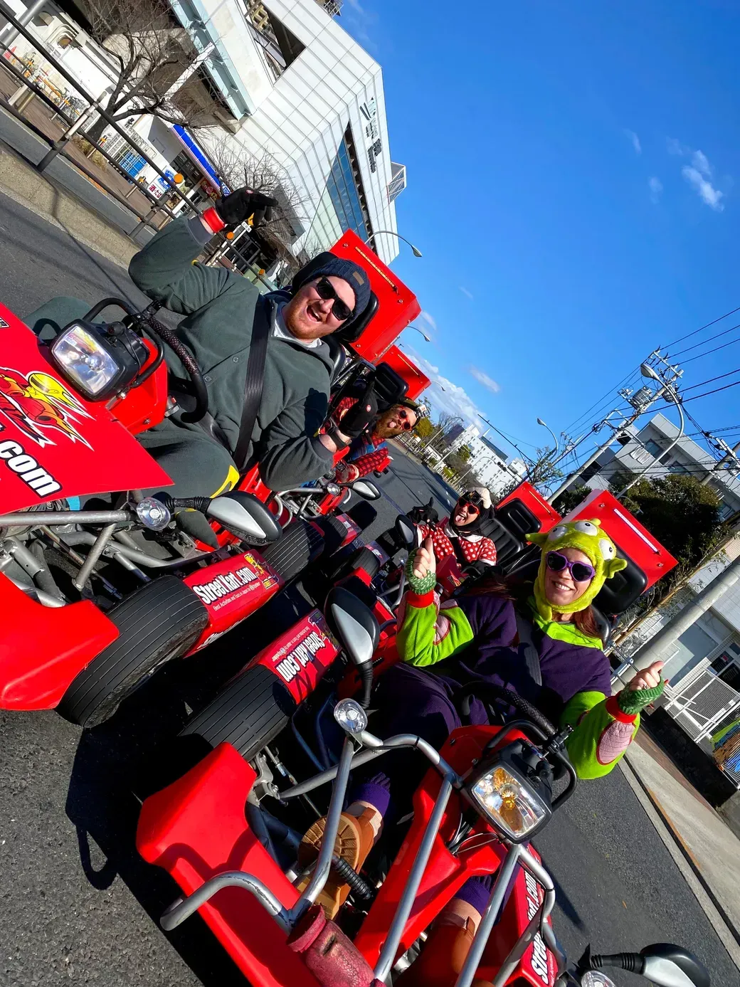 Three people in colorful costumes ride bright red go-karts on a sunny city street, giving thumbs up.
