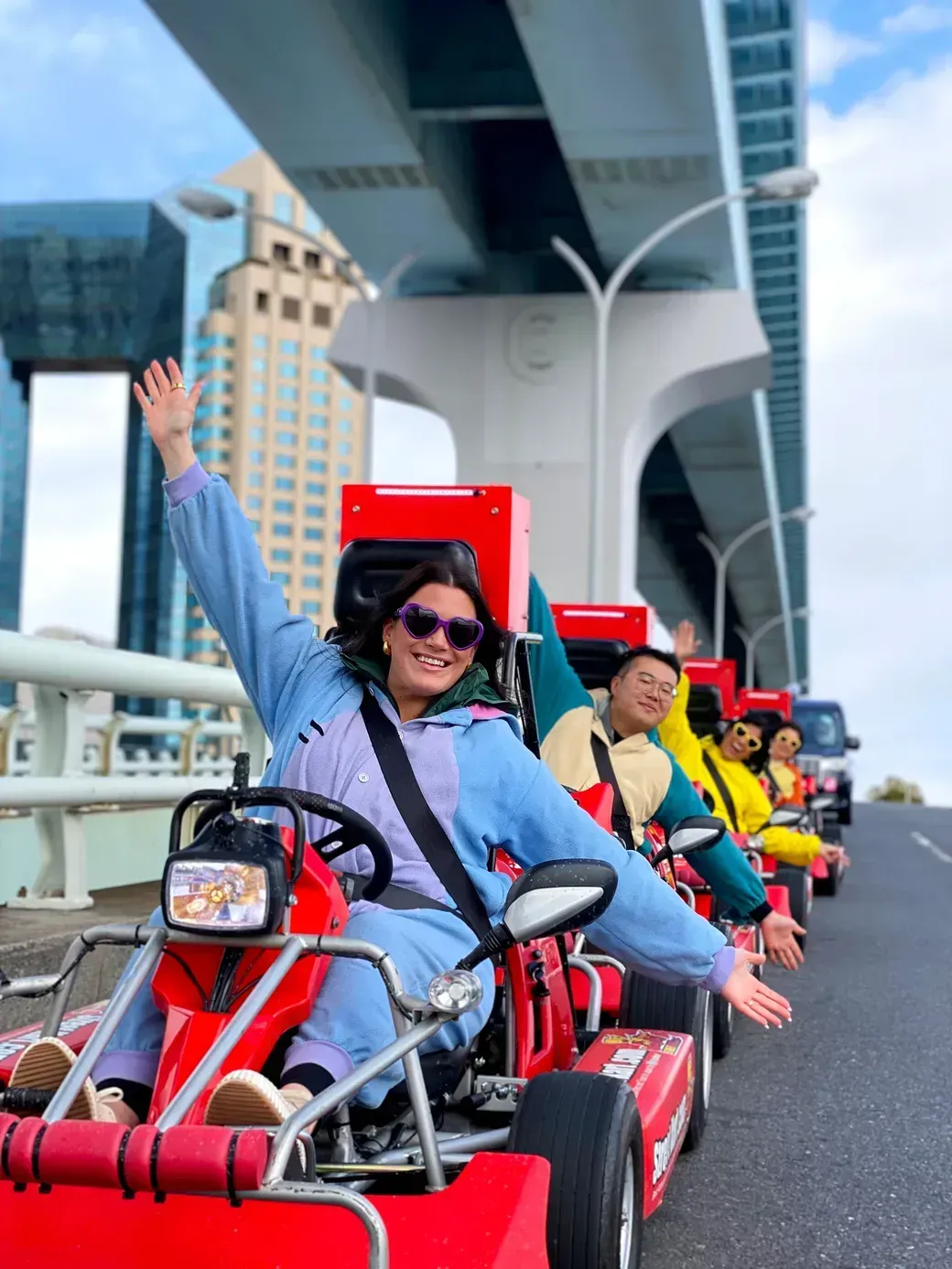 Group of people riding red go-karts along a city street under a large elevated bridge, smiling and waving at the camera.