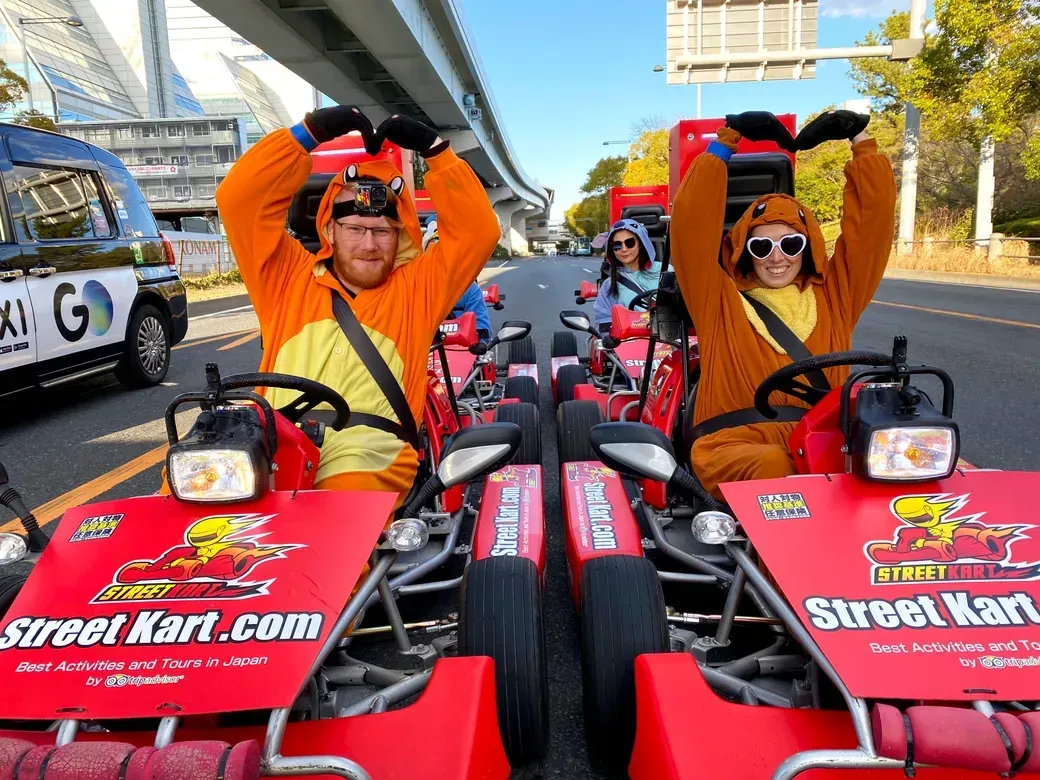 Two people in orange mascot outfits sit in red Street Kart go-karts on a city street, forming heart shapes with their arms.