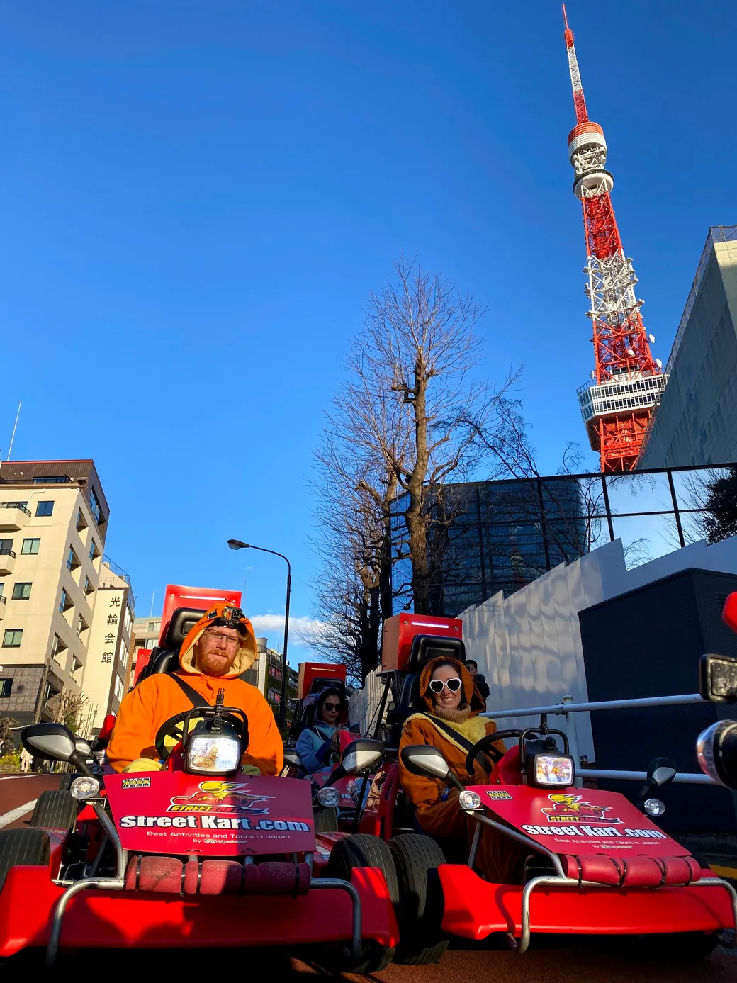 People in orange costumes drive red go-karts labeled StreetKart.com along a city street with Tokyo Tower in the background and a clear blue sky.